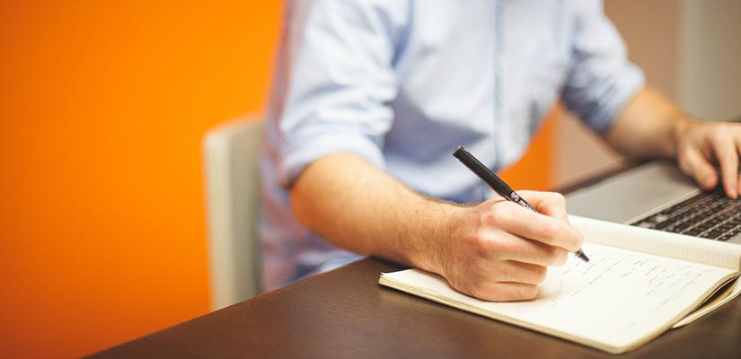 Man sitter i orange konferensrum och skriver vid ett skrivbord. På bordet ligger papper och han håller i en penna.